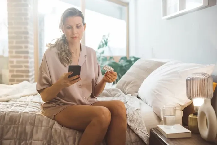 A woman sitting on a bed holding pills and looking at her cell phone.